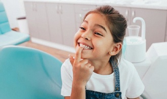 little girl pointing to her smile in dentist’s office 