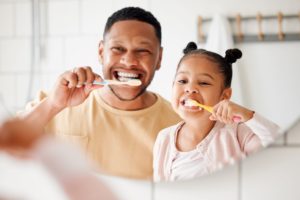 Man in tan shirt and little girl in pink shirt brushing their teeth together in a bathroom mirror